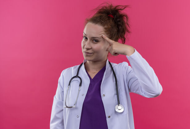 穿着年轻的女医生穿着白大褂 带着听诊器 自信地指着她的太阳穴 站在粉色的墙上专注于任务专注指点女人