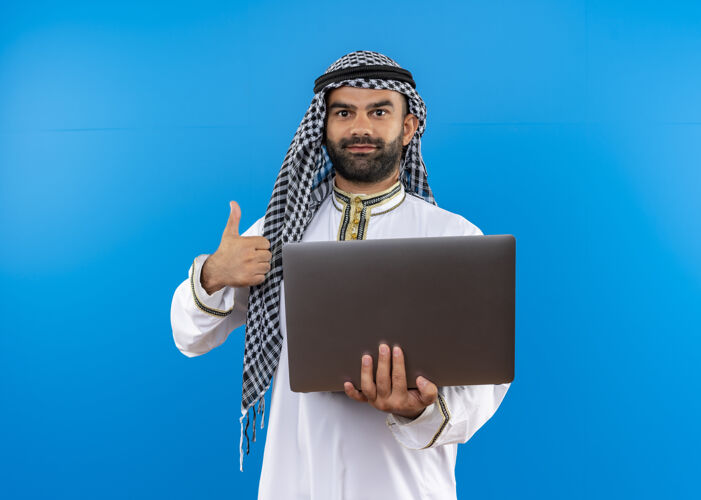 拿着身着传统服装的阿拉伯商人手持笔记本电脑微笑着竖起大拇指站在蓝色的墙上微笑显示穿