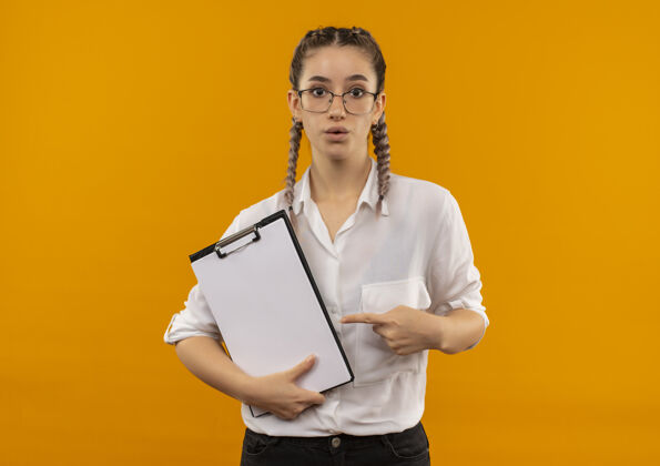 年轻人戴着眼镜 梳着辫子 穿着白衬衫的年轻女学生拿着写字板 用手指着写字板 站在橙色的墙上 看上去很担心女孩人学生