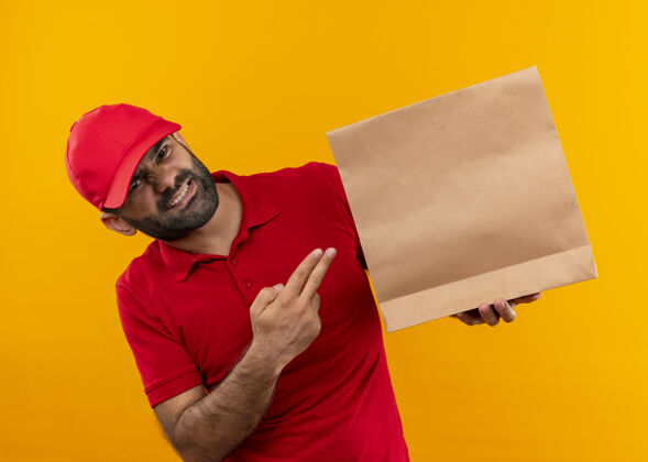表情一个留着胡须 身穿红色制服 戴着帽子的送货员站在橙色的墙上 用手指着纸袋 脸上带着恼怒的表情指着看手指