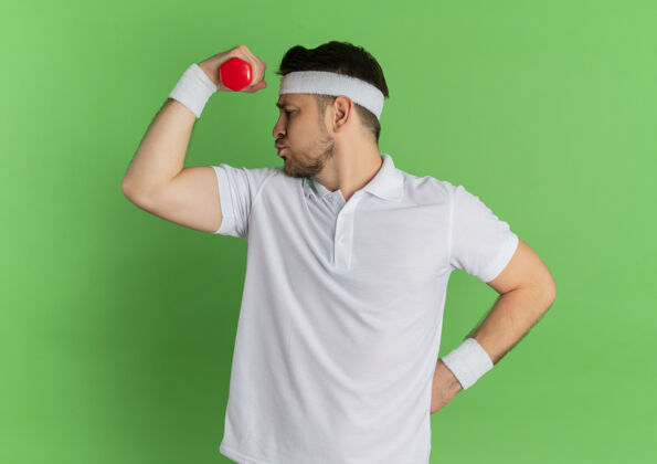 健康身穿白衬衫 头戴钢带的年轻健身男子 带着哑铃 紧张而自信地站在绿色的墙上男性工作健身