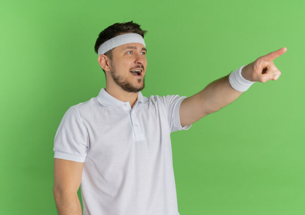 运动装一个身穿白衬衫 戴着头巾的年轻健身男子 用手指着绿色墙壁上站着的东西 愉快地微笑着头带男人微笑