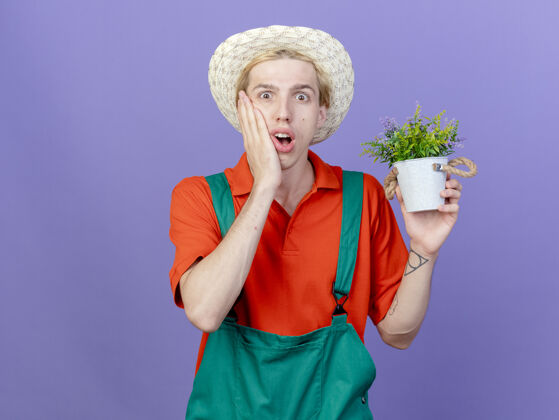 年轻年轻的园丁穿着连体衣 戴着帽子 手里拿着盆栽植物 感到震惊连身衣男人是