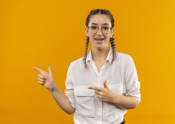 女孩戴着眼镜 梳着辫子 穿着白衬衫的年轻女学生微笑着看着前面 用食指指着站在橙色墙上的一边微笑目录站