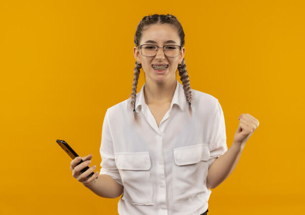 学生戴着眼镜 梳着辫子 穿着白衬衫 手持智能手机 握紧拳头的年轻女学生站在橘色的墙上 快乐而兴奋手势手机人