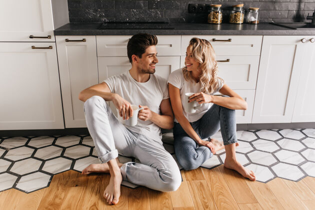 咖啡穿着牛仔裤的可爱女孩坐在地板上和男朋友聊天年轻夫妇在厨房里喝咖啡积极金发室内