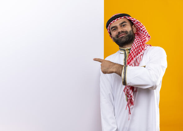 脸身着传统服装的阿拉伯商人站在空白的广告牌旁 手指指着广告牌 脸上带着自信的微笑 越过橙色的墙壁手指传统信心