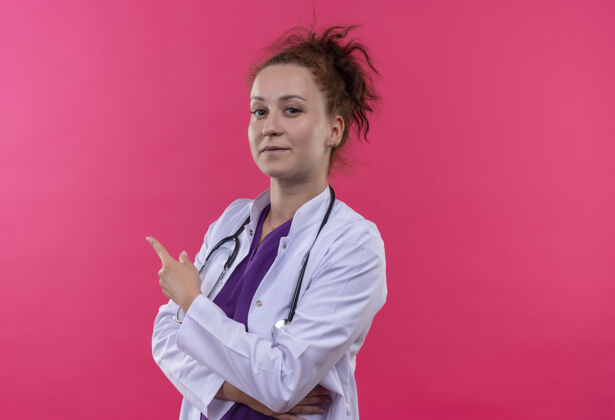 指点年轻的女医生穿着白大褂 带着听诊器 自信地用手指着站在粉红色墙上的一边外套站穿