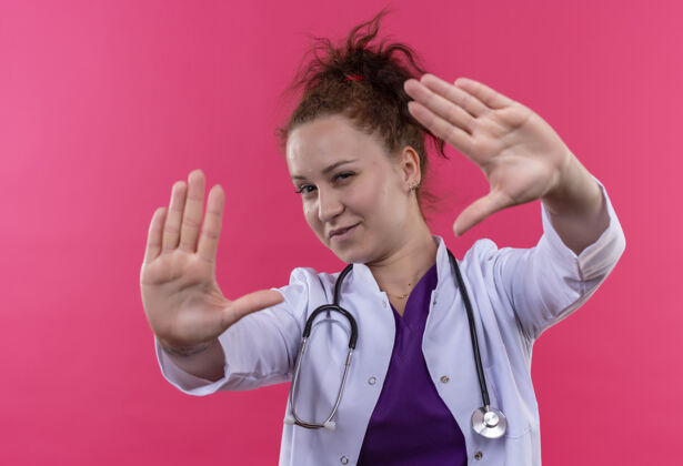 女人年轻的女医生身穿白色外套 手持听诊器 双臂抱着自信的表情站在粉色的墙上手臂听诊器制造