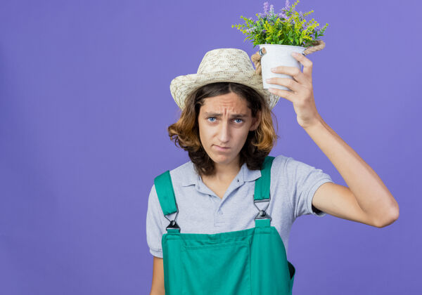 男人年轻的园丁 穿着连体衣 戴着帽子 头上拿着盆栽植物穿抱着帽子