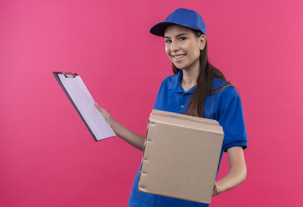 送货年轻的送货女孩穿着蓝色制服 戴着帽子 拿着盒子和剪贴板 看上去自信地微笑着站着帽子粉色