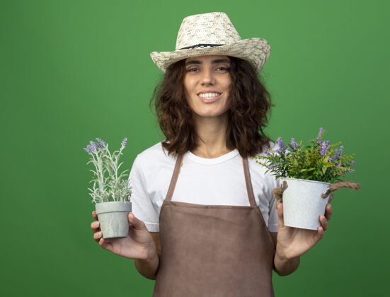 年轻微笑着的年轻女园丁 穿着制服 戴着园艺帽 把花盆里的花隔离在绿色的墙上花抱着花园