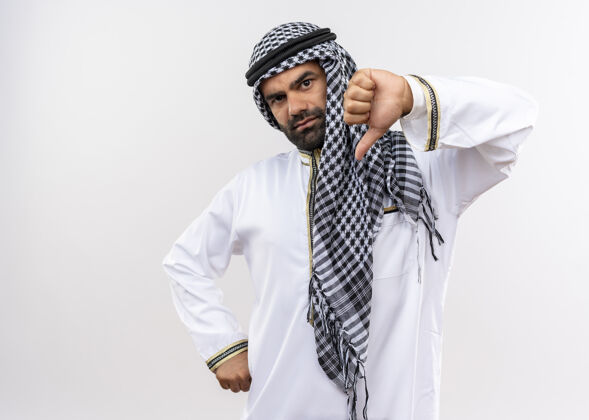 不高兴一个穿着传统服装的阿拉伯男人站在白色的墙上竖起大拇指 很不高兴阿拉伯语传统拇指