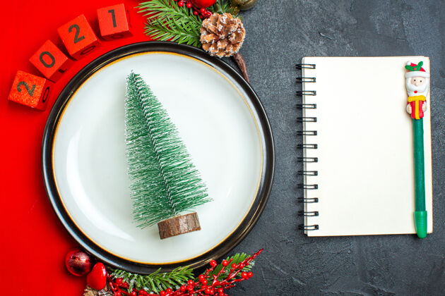餐巾新年背景俯视图 圣诞树晚餐盘装饰配件杉木树枝和数字放在一张红色餐巾上 笔记本旁边是一张黑色桌子上的钢笔树枝容器笔记本