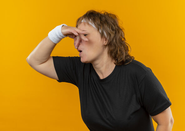 运动中年运动女性 身穿黑色t恤 戴着头巾 手指贴着鼻子站在橙色的墙上 忍受着恶臭痛苦封闭站着