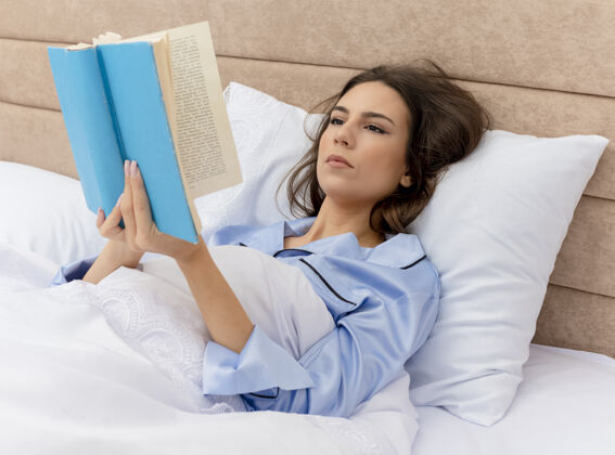躺着穿着蓝色睡衣躺在床上的年轻漂亮女人阅读美丽睡衣