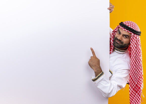 穿身着传统服装的阿拉伯商人站在橙色的墙上 用手指指着空白的广告牌 微笑着看着它空白微笑传统