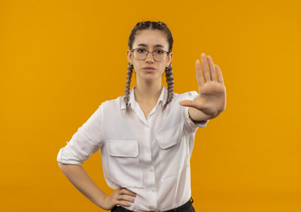 站戴着眼镜 梳着辫子 穿着白衬衫的年轻女学生 手朝前看 严肃的脸站在橙色的墙上脸摆姿势站