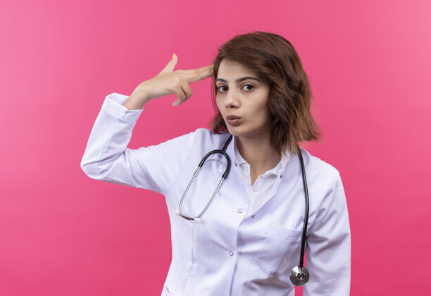 身穿白大褂 手持听诊器的年轻女医生站在粉色墙壁上 在太阳穴附近做手指或枪的手势做站着枪
