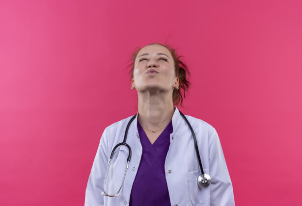 女人年轻的女医生穿着白大褂 带着听诊器 站在粉红色的墙上 高兴而积极地抬头看年轻阳性穿