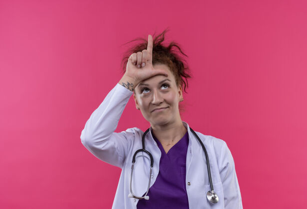 站年轻的女医生穿着白大褂 手持听诊器 站在粉红色的墙上 头上举着失败者的牌子头医生穿