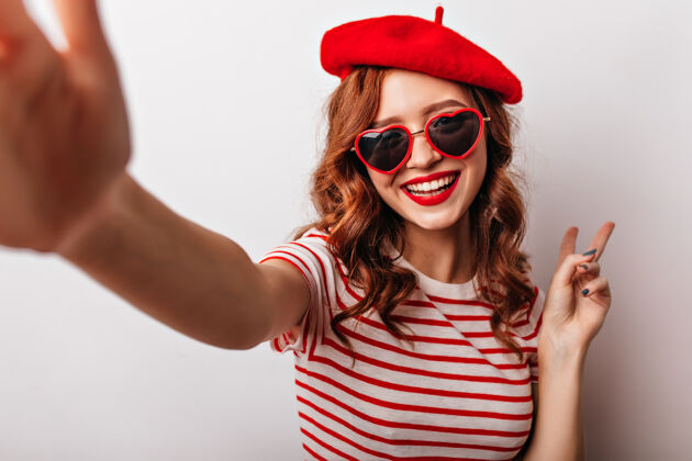 法国幸福的法国女人戴着太阳镜自拍热情的卷发红发女孩在白墙上摆姿势帽子卷发深色