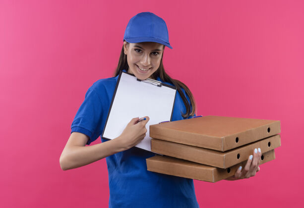 粉色穿着蓝色制服 戴着帽子的年轻送货女孩手里拿着一叠披萨盒和空白页的剪贴板 要求签名帽子年轻页面