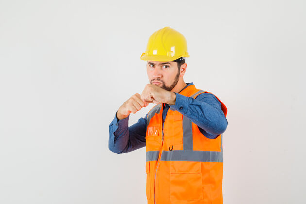 工人年轻的建筑工人站在拳击手的姿势在衬衫 背心 头盔 并期待着认真前视图硬工厂工业