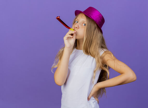 生日美丽的小女孩在节日帽子吹口哨看着相机 生日派对的概念站在紫色的背景下女孩看派对