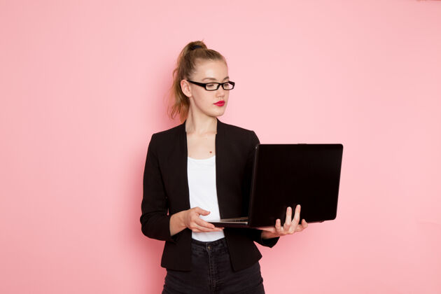 前面身穿黑色夹克的女办公室工作人员正拿着笔记本电脑在浅粉色的墙上使用工人专业人