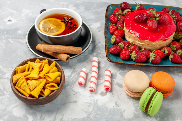 饼干正面近距离观看美味的草莓蛋糕与新鲜的红色草莓一杯茶和麦卡龙在白色表面美味封闭盘子