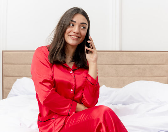 微笑穿着红色睡衣的年轻漂亮女人坐在床上微笑着 在卧室内部的灯光背景下讲手机女人红色睡衣