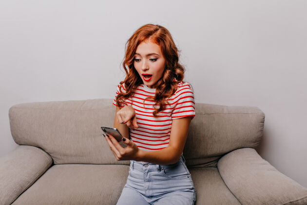 室内坐在沙发上惊讶地看着手机屏幕的女士穿着条纹t恤的姜汁女孩和智能手机合影红发漂亮情感