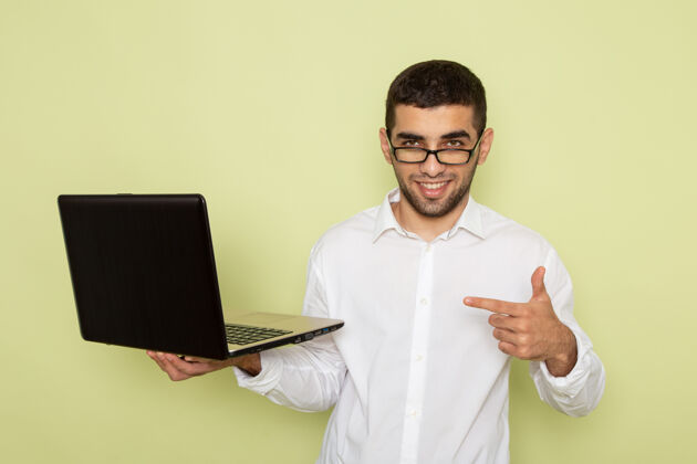 个人身穿白衬衫的男性上班族手持笔记本电脑 微笑着站在浅绿色的墙上男性电脑专业人士