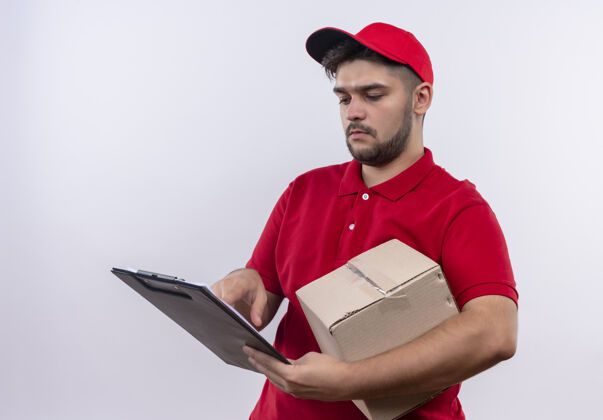 空白年轻的送货员穿着红色制服 戴着帽子 手里拿着一个盒子和一张白纸的剪贴板 严肃地看着他们包装盒子递送