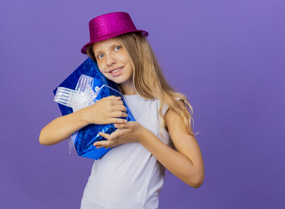 帽子戴着节日礼帽的漂亮小女孩抱着礼盒看着相机笑容可掬 紫色背景下站着生日派对的概念快乐礼物聚会