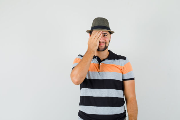 友好一个穿着t恤衫 戴着帽子的年轻人 一只手举着一只眼睛 面带羞耻的表情 正对着前方站立帽子胡茬