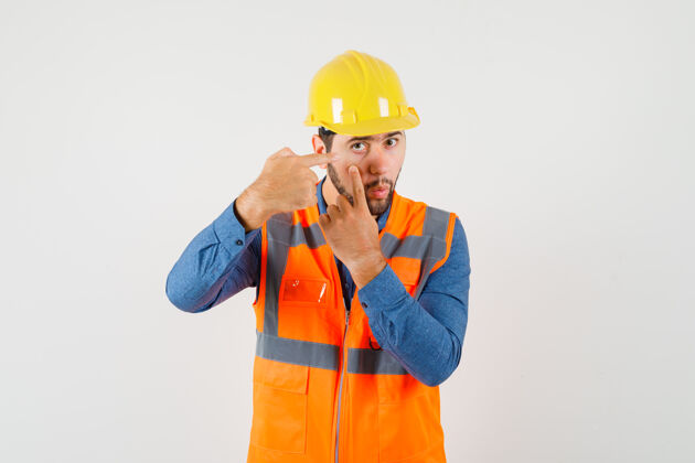 车间年轻的建筑工人穿着衬衫 背心 头盔指着手指拉着的眼皮 前视图帽子工作工业