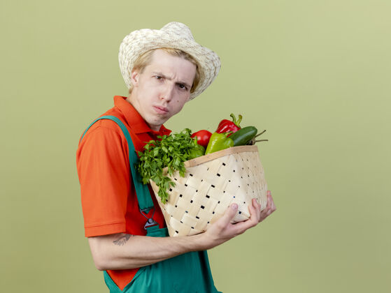 站着年轻的园丁穿着连体衣 戴着帽子 手里拿着装满蔬菜的箱子 站在灯光下严肃地皱着眉头看着摄像机皱眉板条箱男人