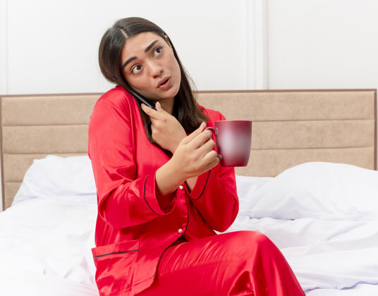女人穿着红色睡衣的年轻漂亮女人坐在床上 端着一杯咖啡讲着手机 在灯光的背景下 卧室里一副迷茫的样子杯子咖啡室内