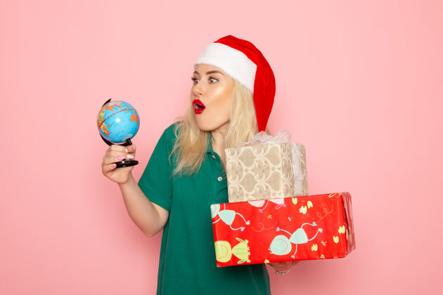 年轻的女性正面图年轻女性手持地球仪和粉色墙上的圣诞礼物照片模特女性圣诞节新年彩色假期风景圣诞节地球仪