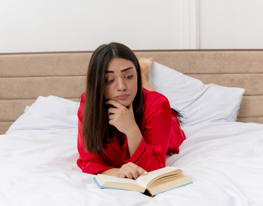 迷茫穿着红色睡衣的年轻美女躺在床上看书 在卧室的室内灯光背景下显得很困惑看床女人