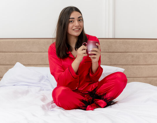 年轻穿着红色睡衣的年轻美女坐在床上 端着一杯咖啡 面带微笑地看着相机 在灯光背景下享受卧室的时光女人微笑看着