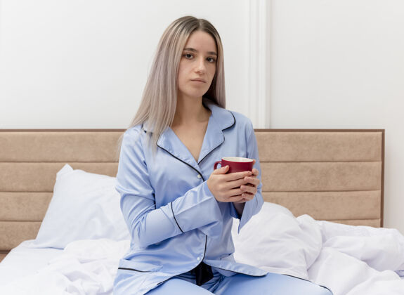 坐着穿着蓝色睡衣的年轻美女坐在床上 端着一杯咖啡 严肃地看着摄像机 在卧室的室内灯光背景下脸室内卧室