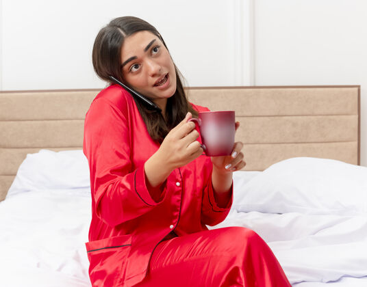床穿着红色睡衣的年轻美女坐在床上 端着一杯咖啡 在卧室的室内灯光背景下用手机聊天手机杯子卧室