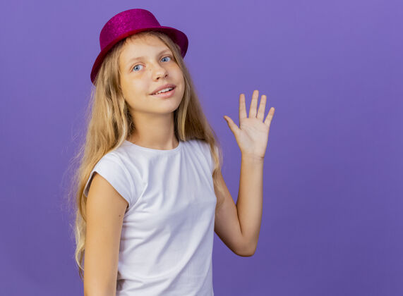 紫色戴着节日礼帽的漂亮小女孩挥手微笑 紫色背景下站着生日派对的概念节日手帽子