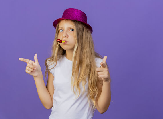手指戴着节日帽子的漂亮小女孩用食指对着镜头吹哨子 紫色背景下站着生日派对的概念小帽子女孩