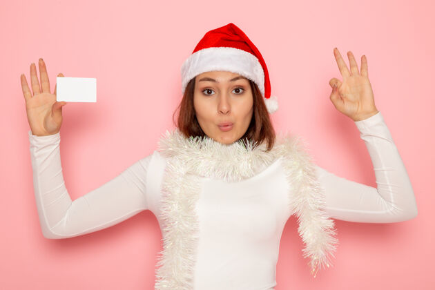 风景正面图年轻女性手持白色银行卡在粉色墙上圣诞假期新年时尚彩钱银行圣诞节帽子