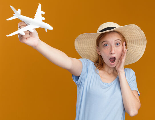 飞机惊讶的年轻红发姜女雀斑戴沙滩帽举行模型飞机孤立在橙色墙上复制空间穿雀斑红发
