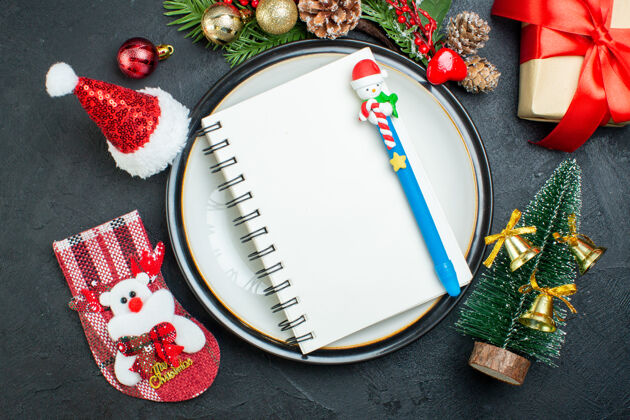 笔记本上图为笔记本 用笔放在餐盘上圣诞树冷杉树枝针叶树圆锥形礼品盒圣诞老人帽子圣诞袜黑色背景盘子圆锥体帽子